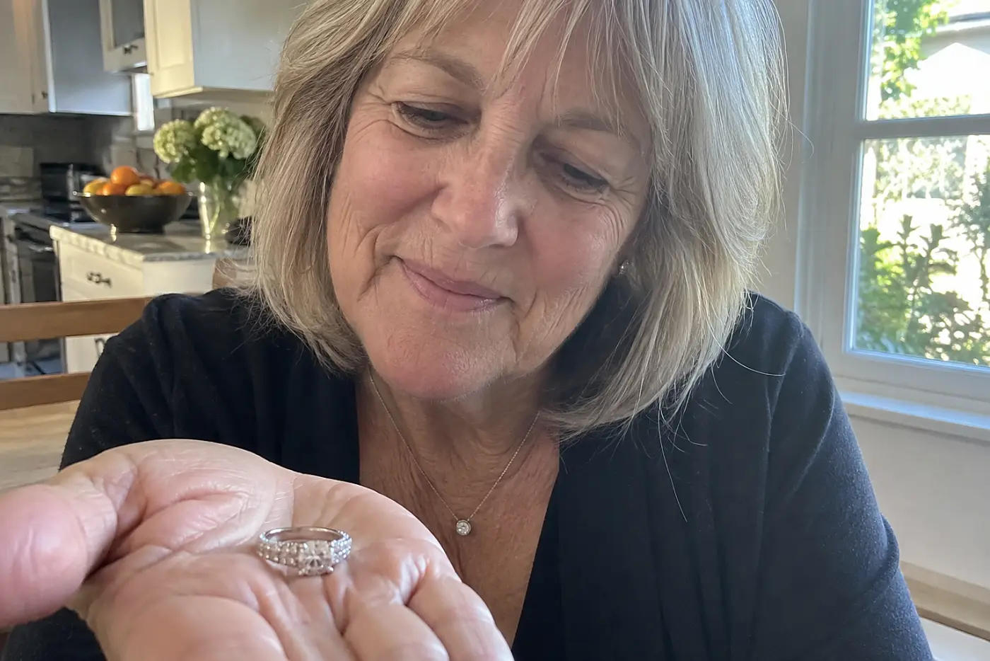 A ring with tiny side diamonds rests in the center of a woman's hand at a kitchen table, with every setting visible.