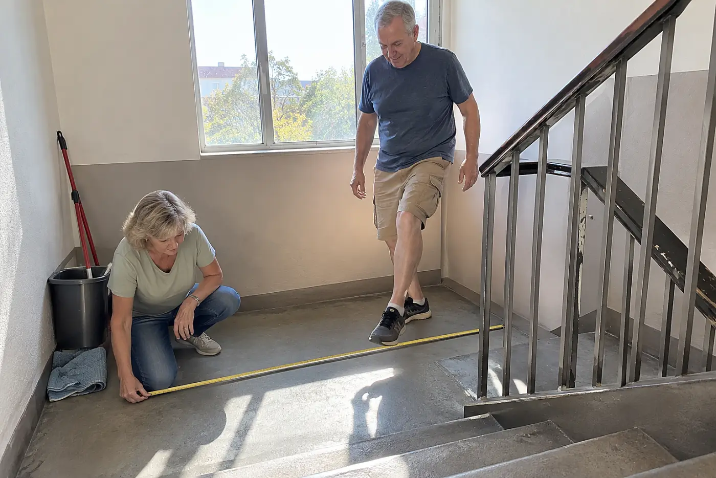 Woman measuring a stair landing beside a railing in an apartment building.