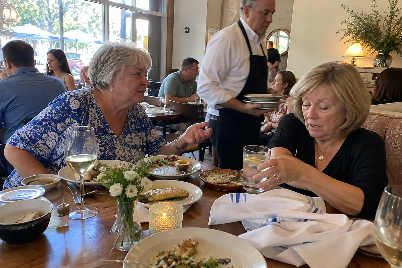 Older woman leaning toward a friend across a busy restaurant table.