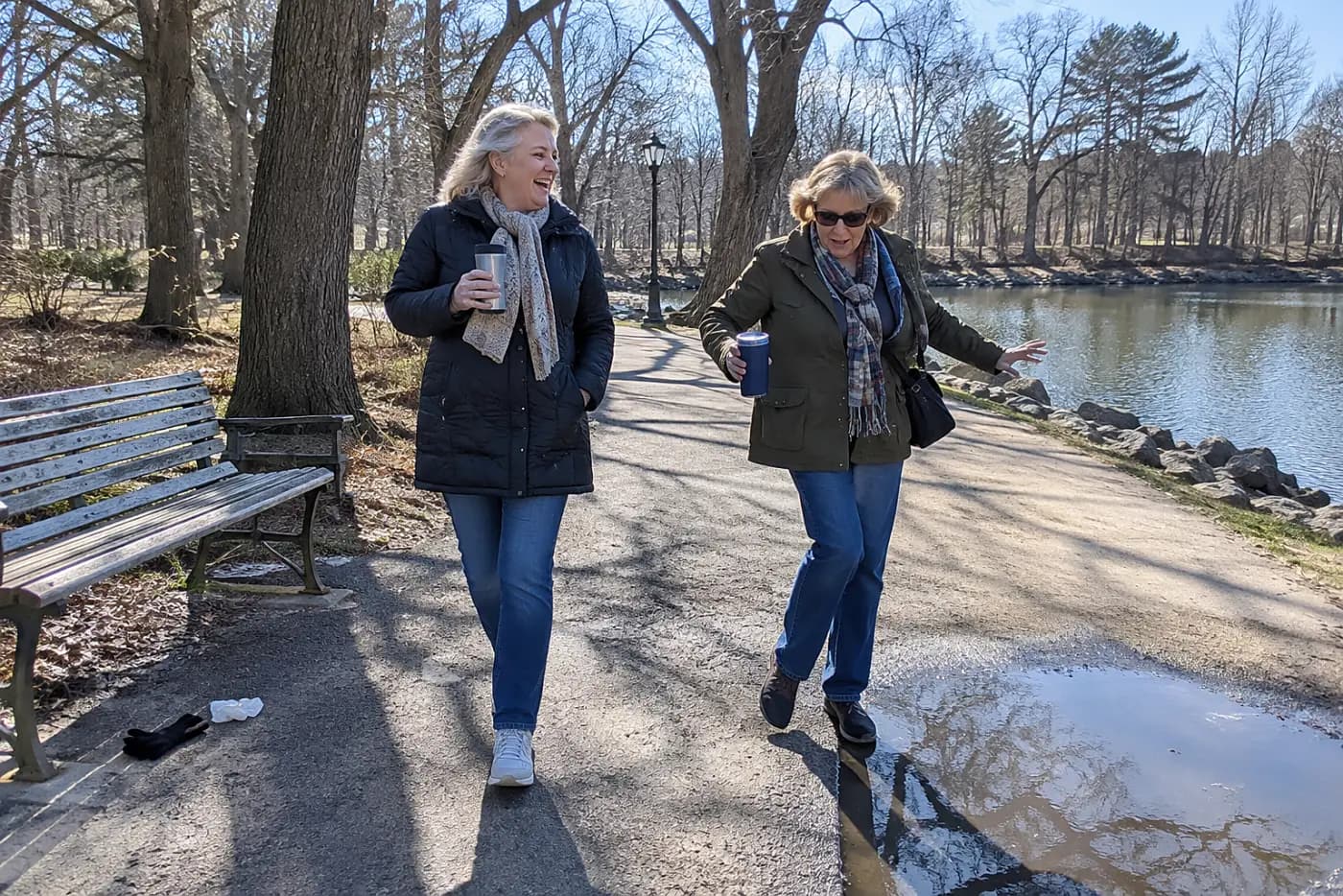 Two older women walking with travel mugs along a park path.