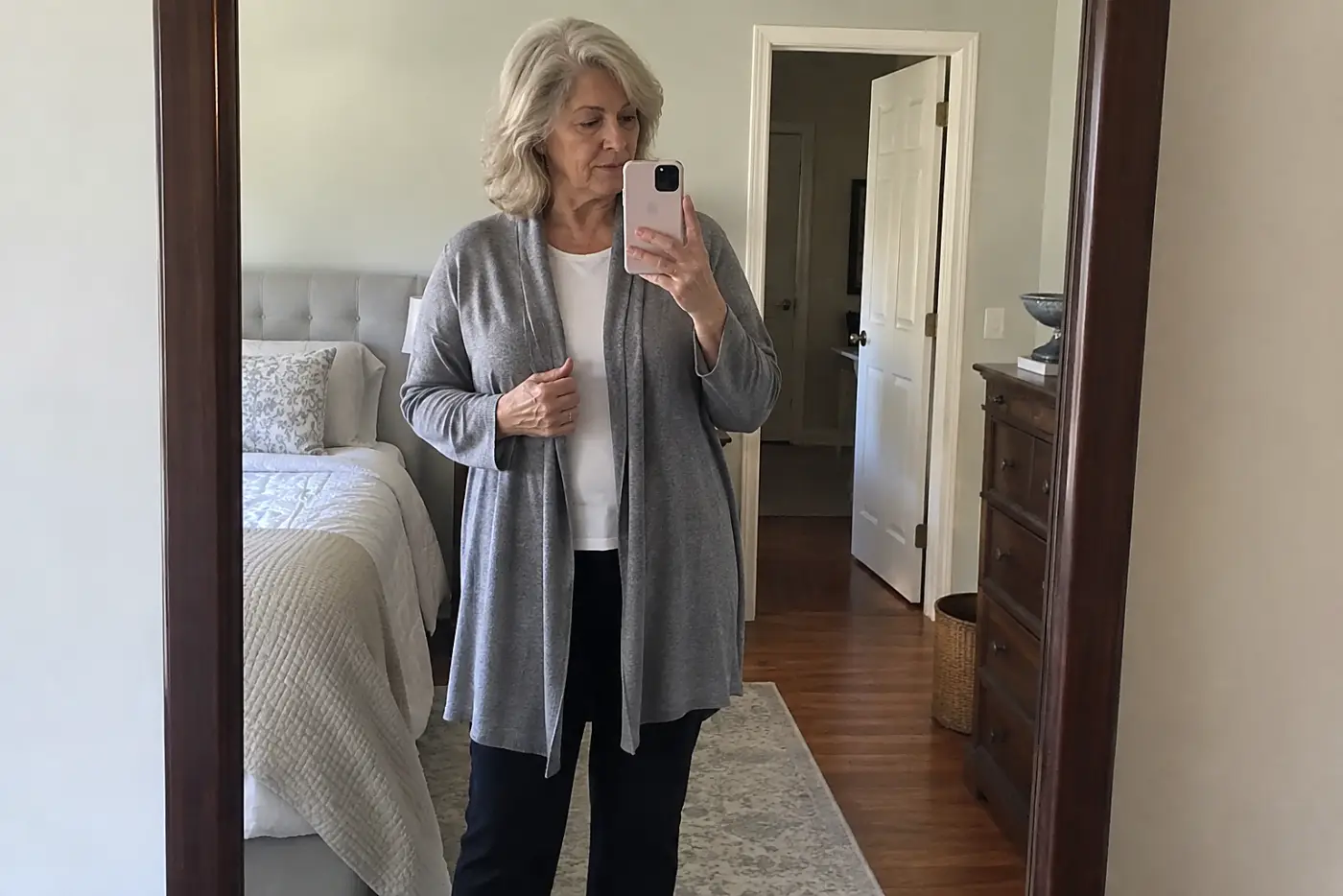 A full-length wall mirror centers a woman in her sixties standing back in her bedroom doorway, where her blouse disappears beneath a long cardigan, with no writing visible.