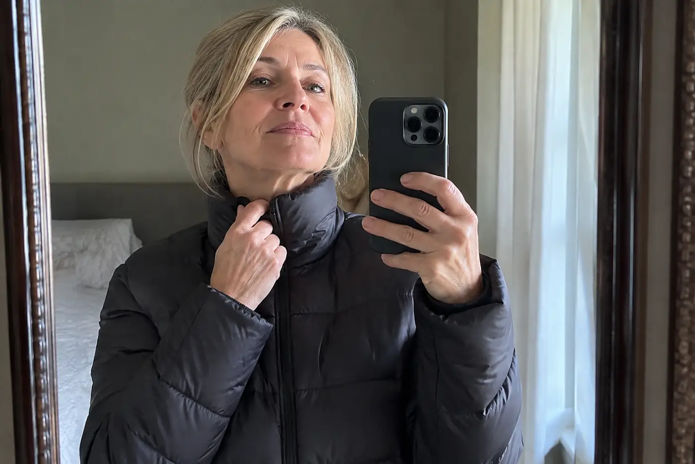A jacket buttoned to the neck fills the frame on a woman over 50 standing before a bedroom mirror, with no writing visible.