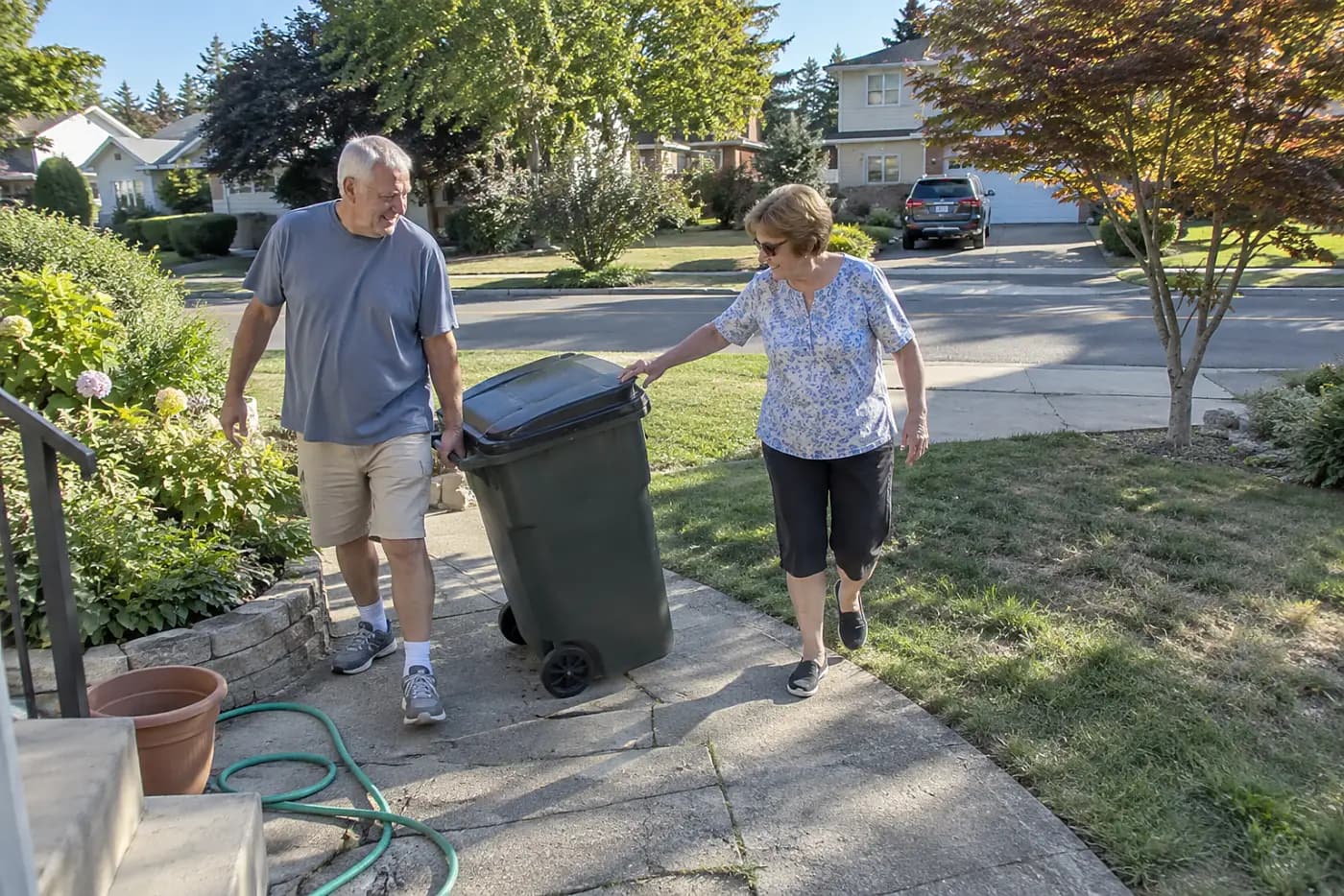 A neighbor carries a trash bin down a front walkway beside an older woman.