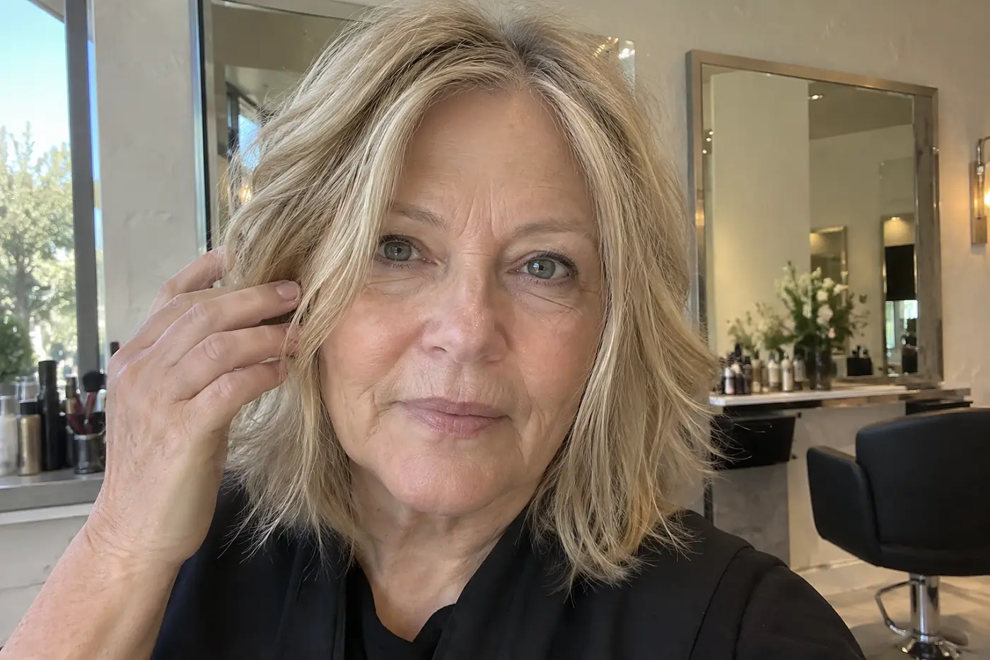 A dry, unstyled face-frame cut is centered on a woman in her sixties checking the front of her hair in a salon mirror by daylight, with no writing visible.