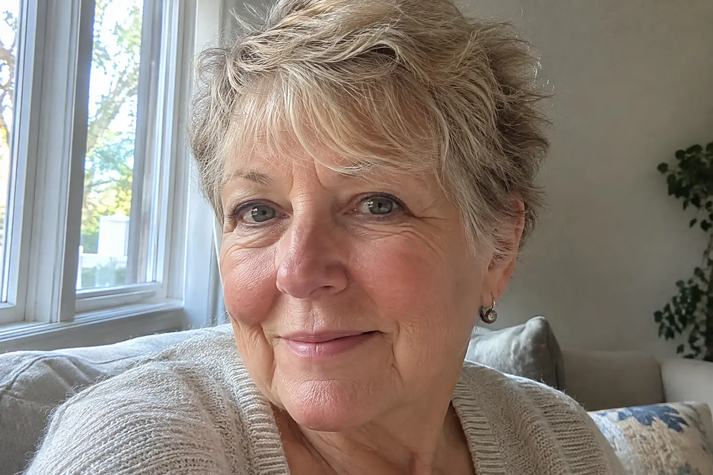 A textured crop with short fringe fills the center of a close portrait of a woman in her sixties near a daylight window, with no writing visible.