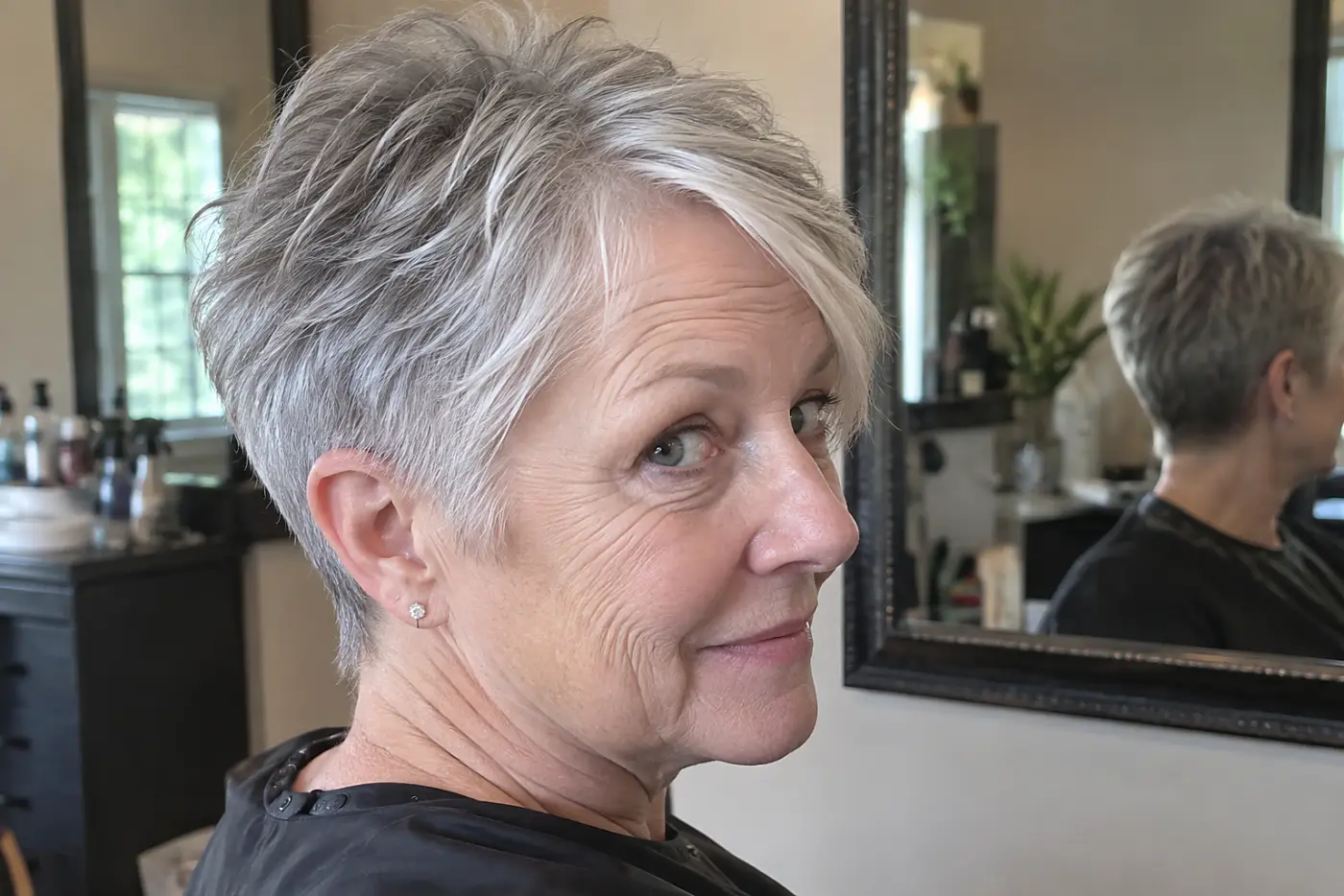 A tapered pixie at the nape fills the center of a side-and-back view of a woman in her sixties in a bright salon, with no writing visible.