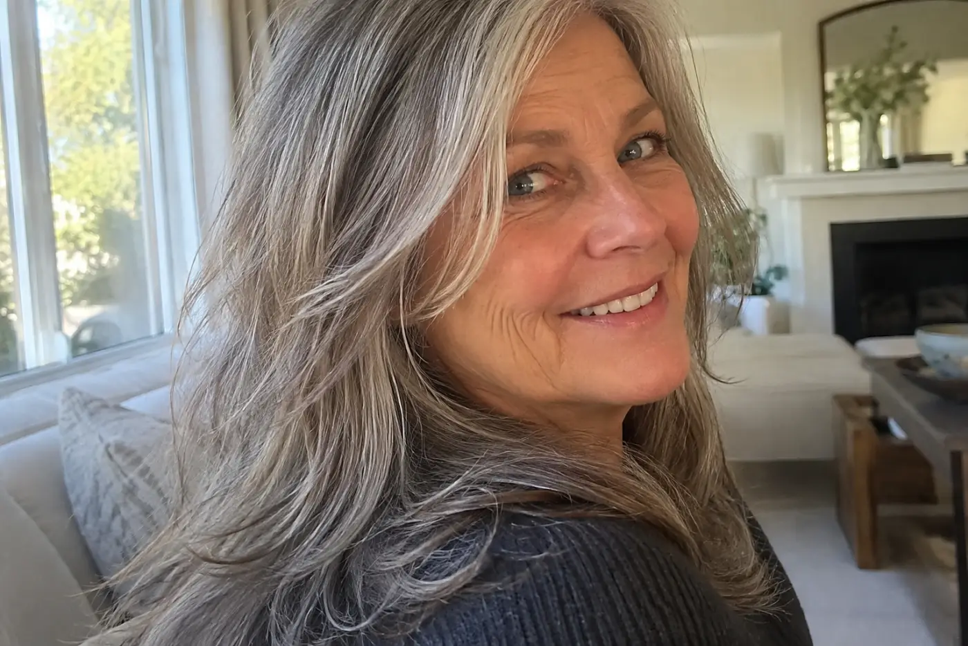 Long layers below the shoulders fill the center of a candid portrait of a woman in her sixties by a living-room window, with no writing visible.