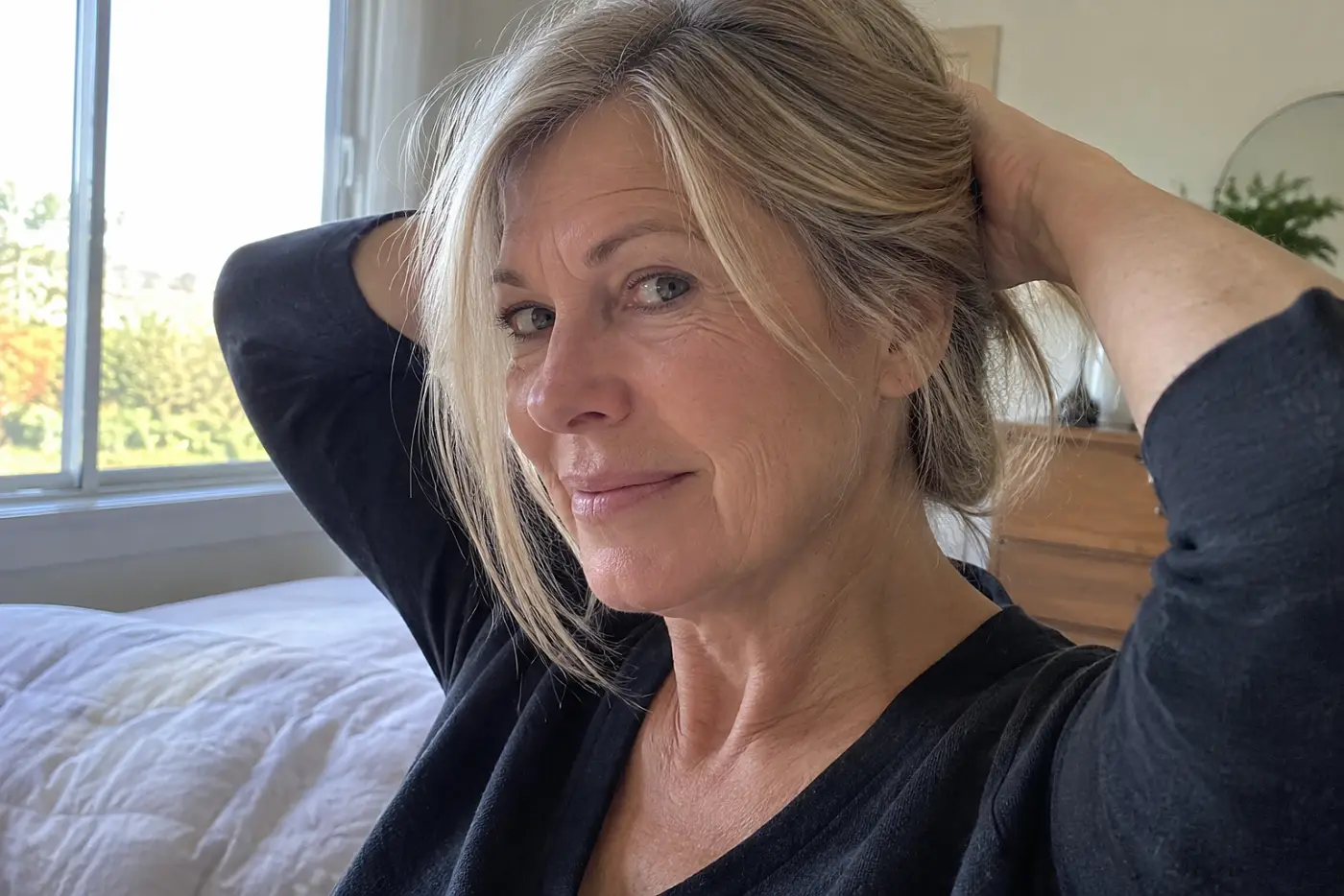 A one-length shoulder cut is centered on a woman in her sixties gathering her hair behind her head in a sunlit bedroom, with no writing visible.