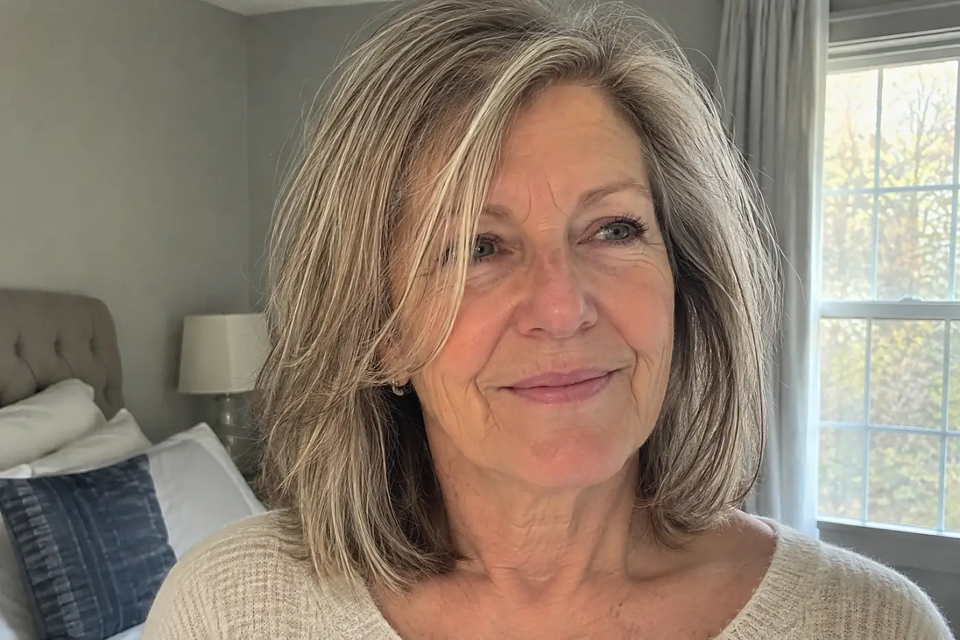 A collarbone-length lob is centered on a woman in her sixties standing by a bright bedroom window, with no writing visible.