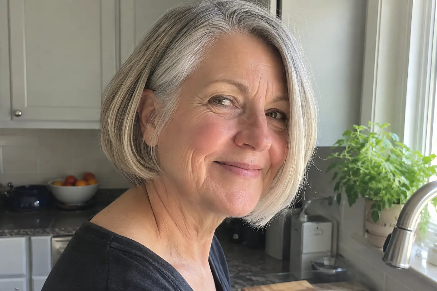 A bob with a side part is centered on a woman in her sixties turning toward daylight in her kitchen, with no writing visible.