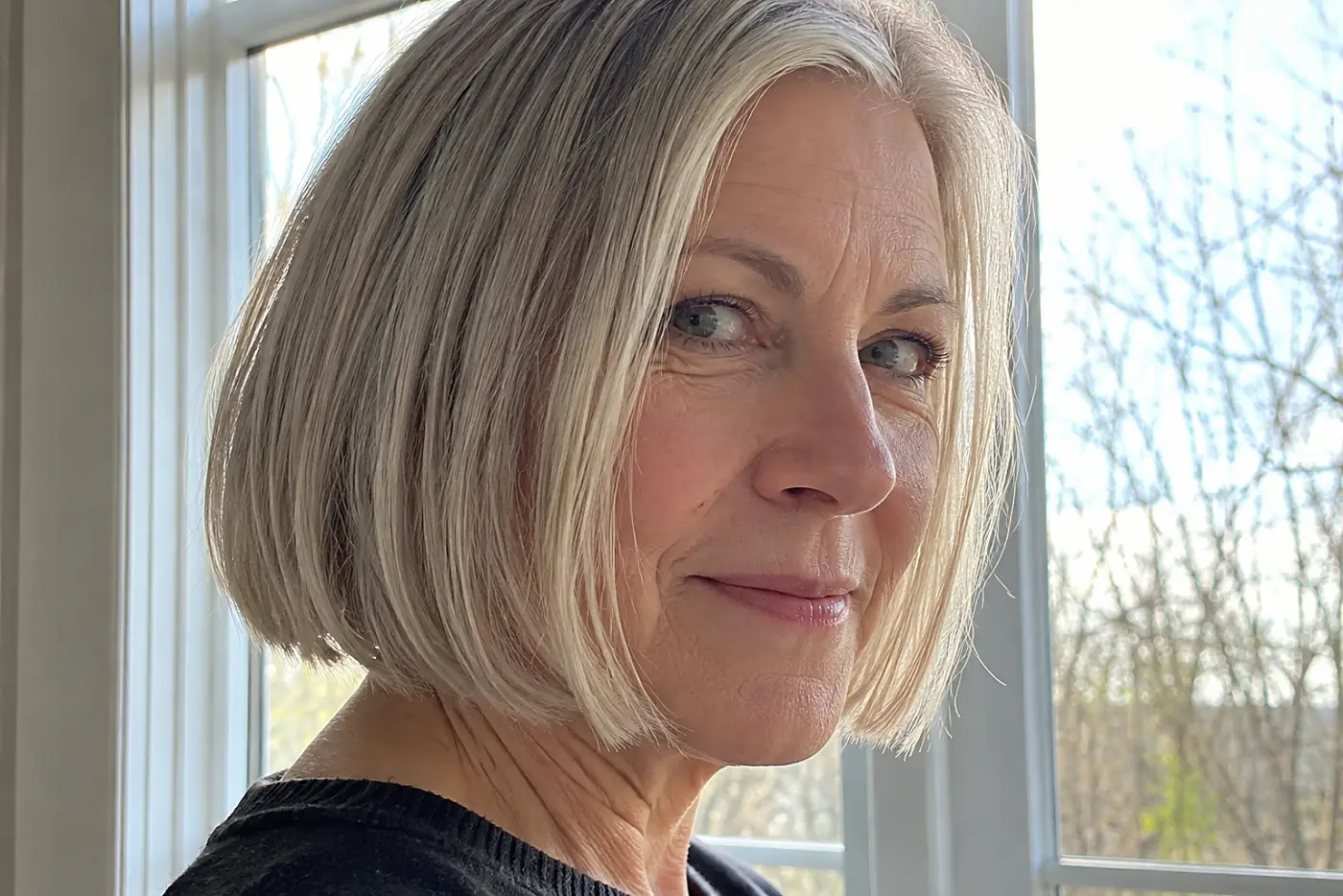 A blunt bob ending at the jaw fills the center of a side-view portrait of a woman in her sixties beside a daylight window, with no writing visible.