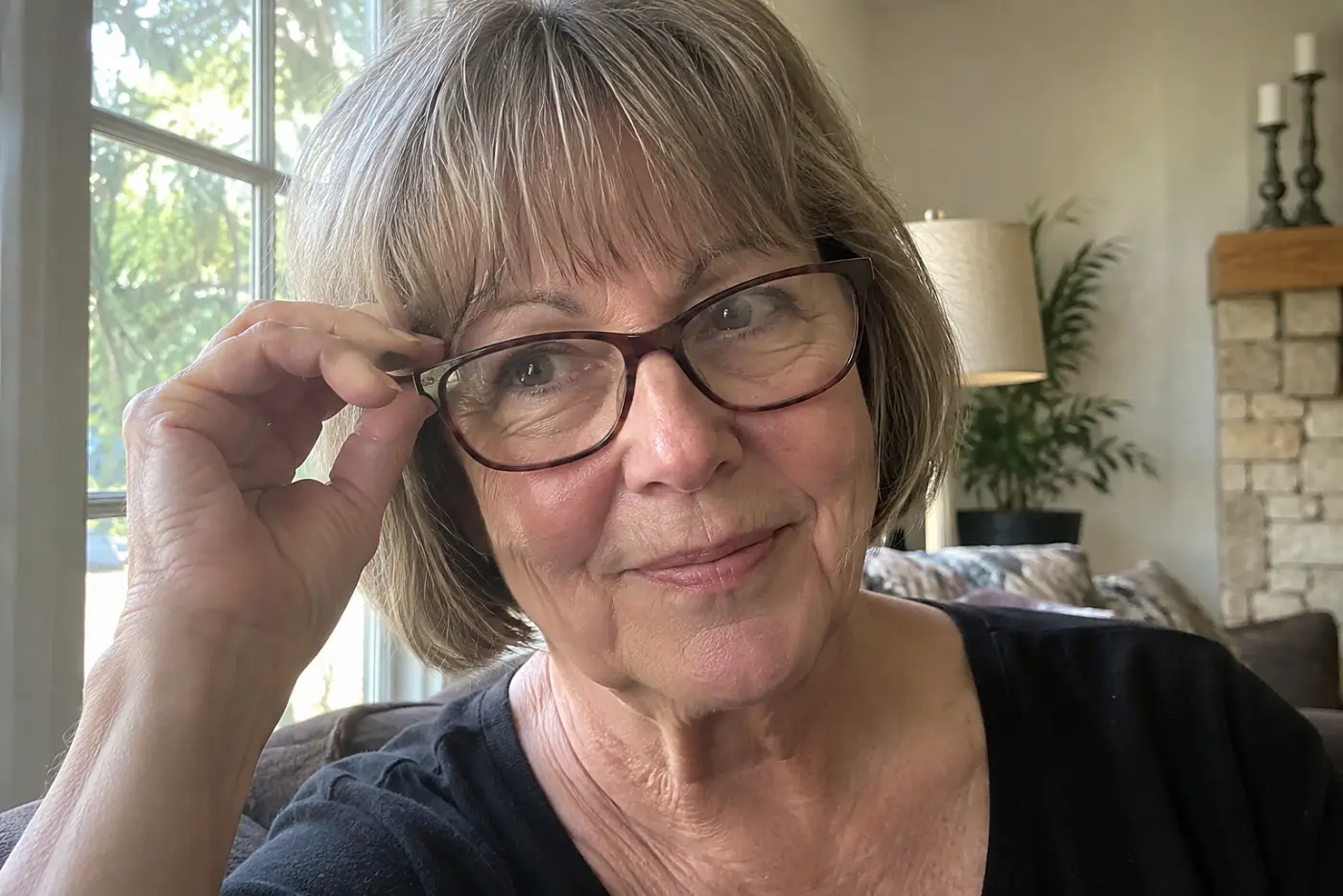 A bob with brow-length bangs is centered on a woman in her sixties wearing plain glasses at home in daylight, with no writing visible.