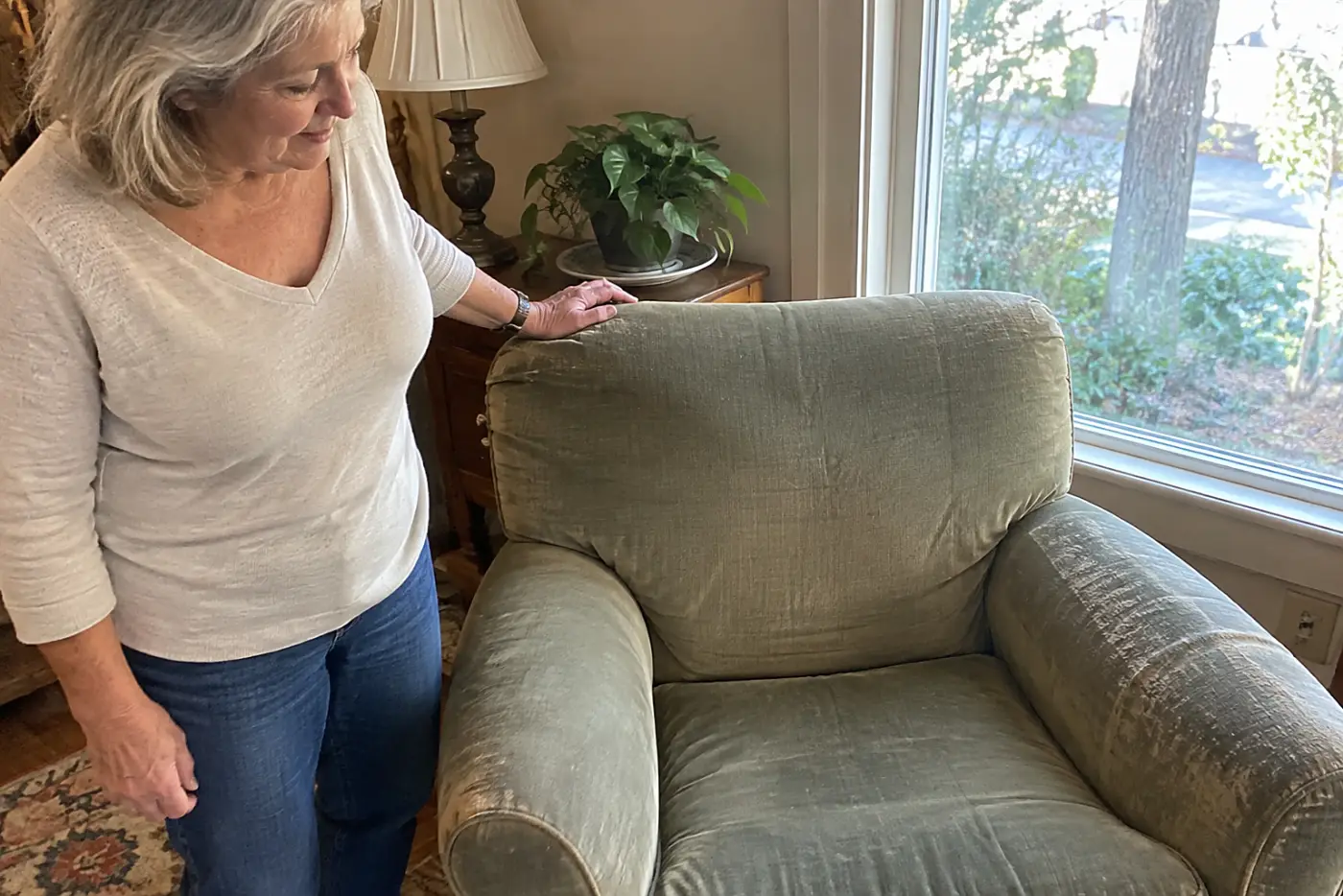 A worn chair stands in the center beside a living-room window as a woman in her sixties rests her hand on its back, with no visible writing.