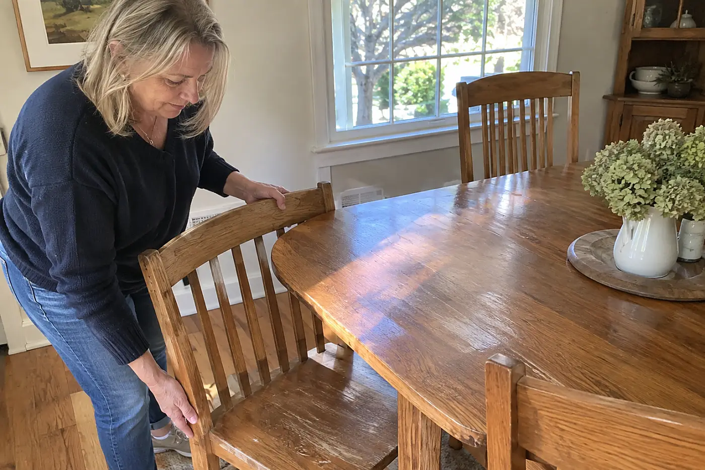 A matching oak dining set fills the center of a family dining room as a woman over 50 pulls out one chair, with no visible writing.