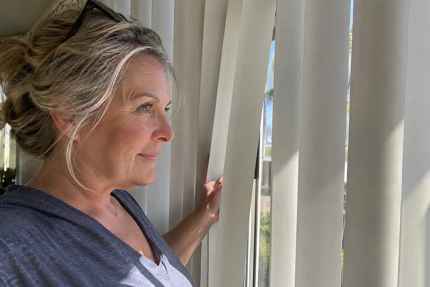 White vertical blinds fill the center across a patio slider as a woman over 50 parts the slats to step outside, with no visible writing.