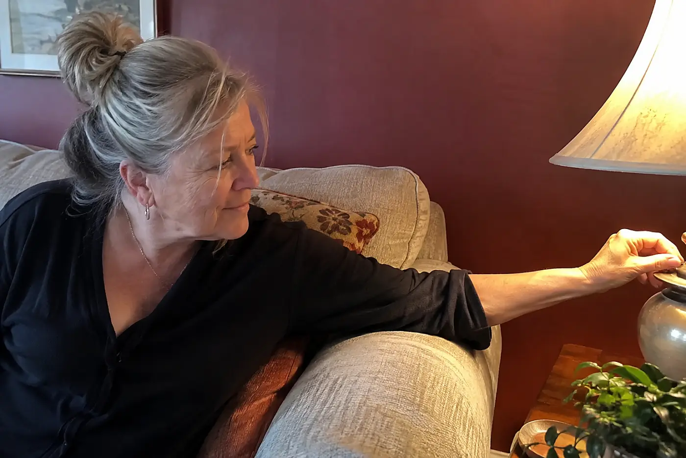 A burgundy accent wall fills the center behind a family-room sofa as a woman over 50 switches on a reading lamp, with no visible writing.