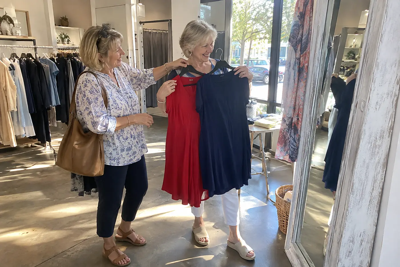 Older woman holding red and navy dresses beside a mirror near a clothing store entrance.