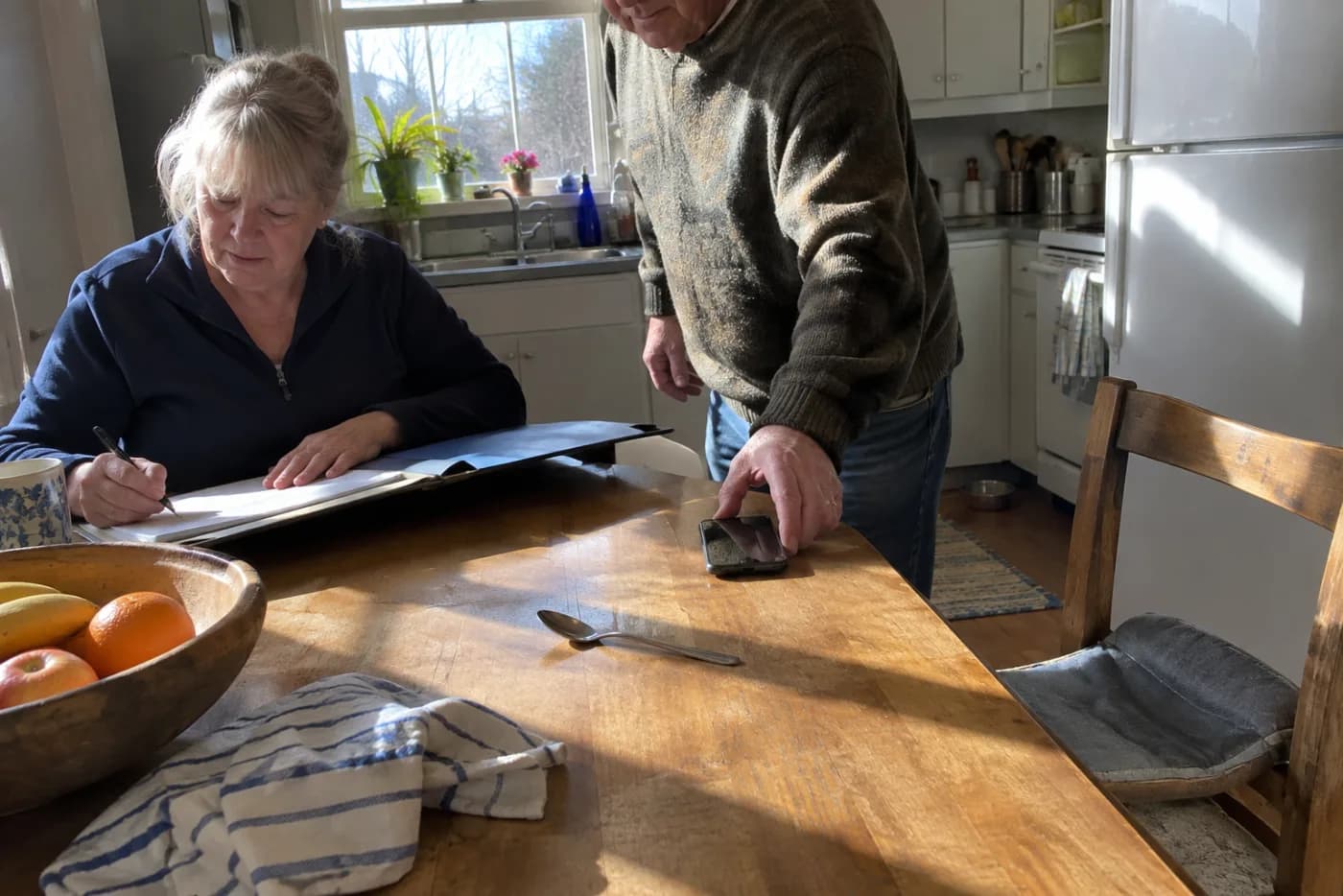 Older woman writing utility account numbers at a kitchen table.