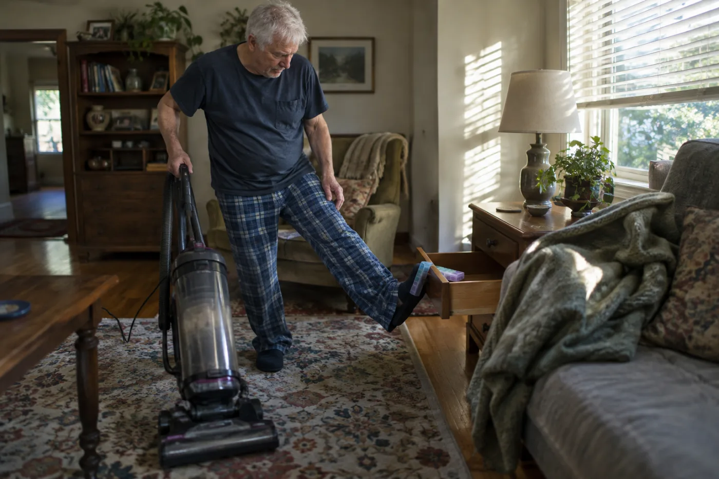 Man vacuuming living room while hiding a pill organizer in a drawer with his foot