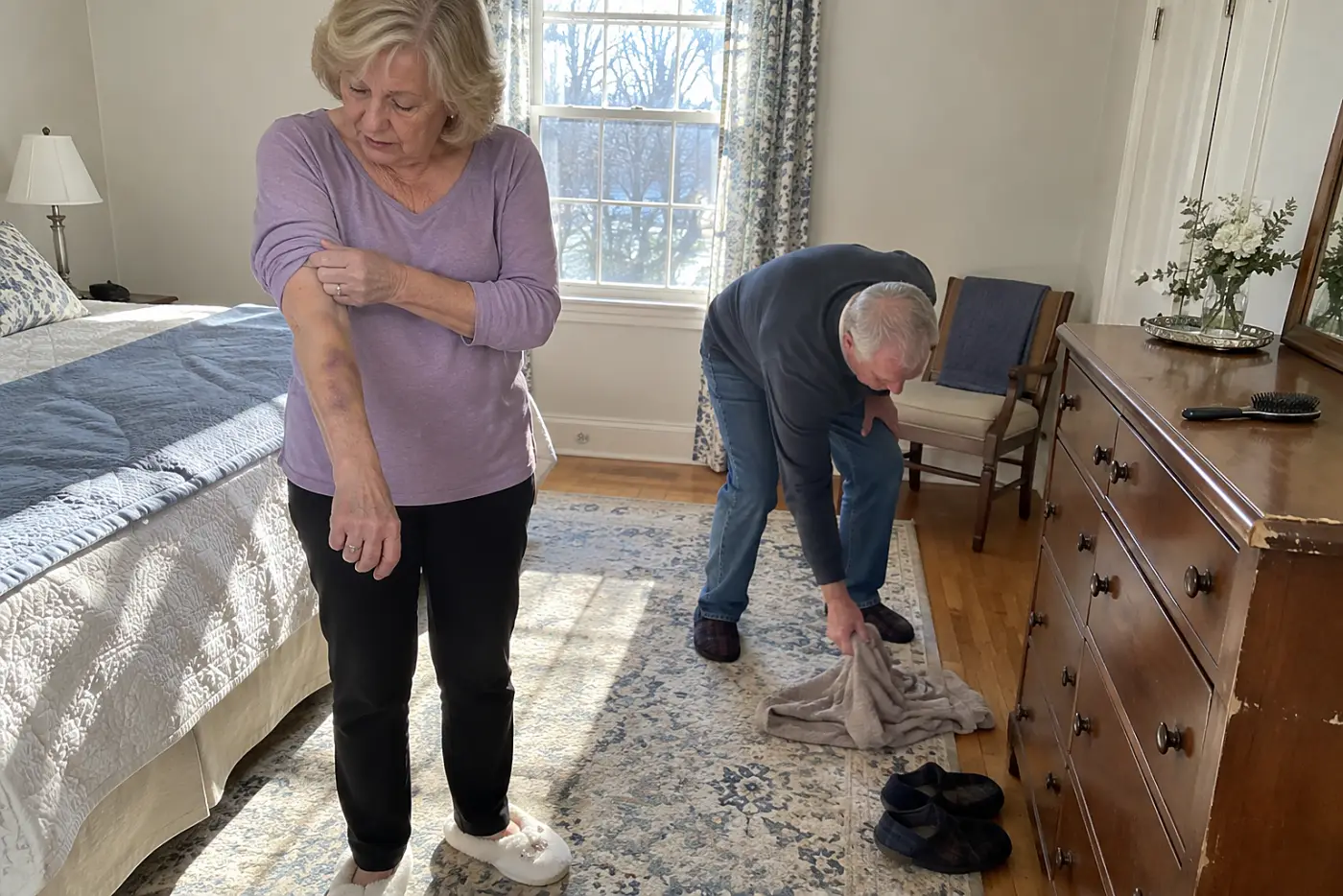 Older woman checking a forearm bruise beside a dresser.