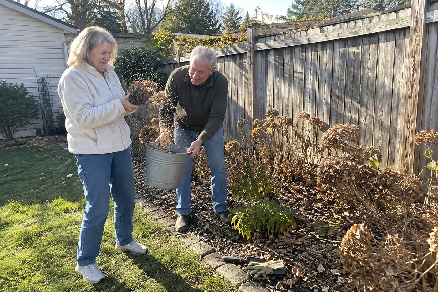 Older woman checking a dry flower head in a small bed by her fence.