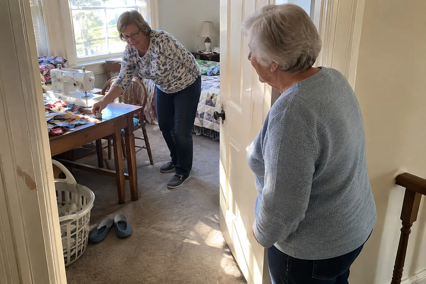 Older woman closing the door to a spare bedroom with a sewing table inside.