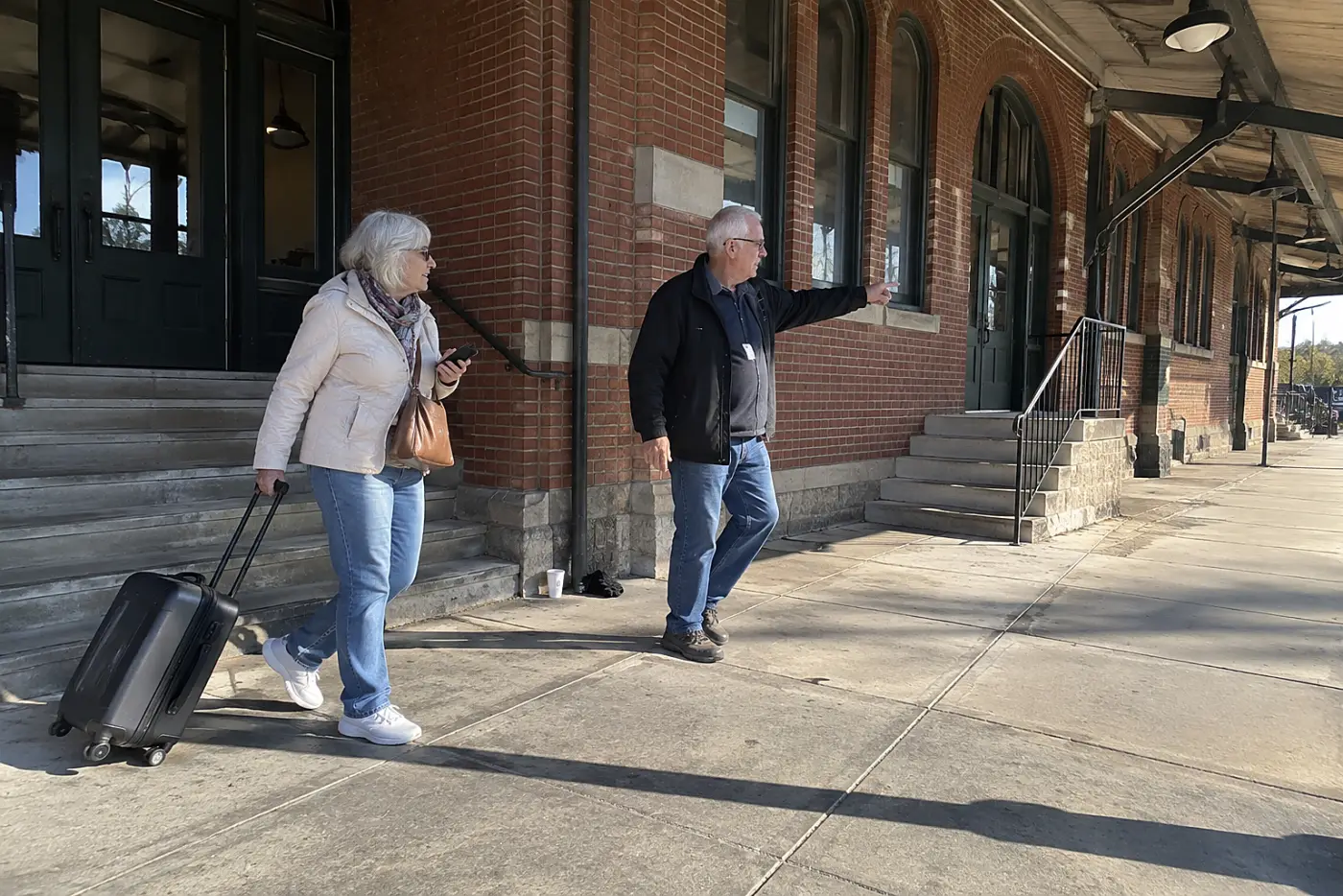 Older woman checking a station entrance on her phone outside a brick train depot.