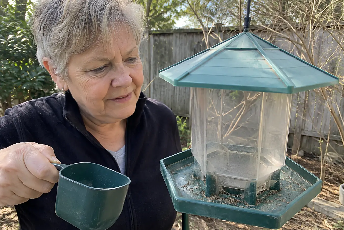 A clean, empty bird feeder hangs in the middle of a backyard above swept ground, with a woman in her sixties holding a small scoop nearby.