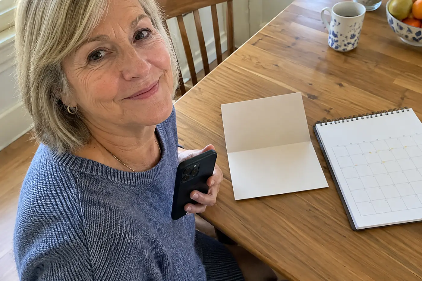 An invitation with no legible writing sits open beside a calendar with blank pages as a woman in her sixties holds her phone.