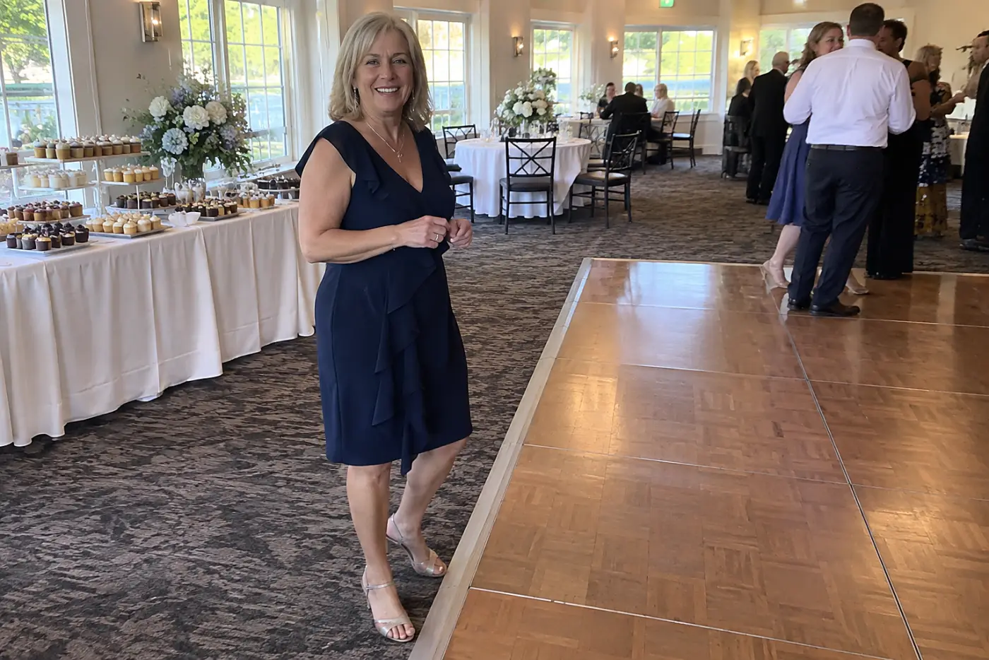 A woman in her sixties stands at the edge of a wedding dance floor near tables set with dessert plates.