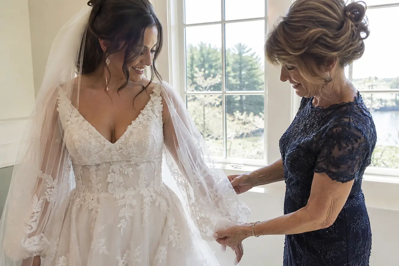 A woman in her sixties admires the bride’s dress beside a window before the ceremony, with both women standing close.