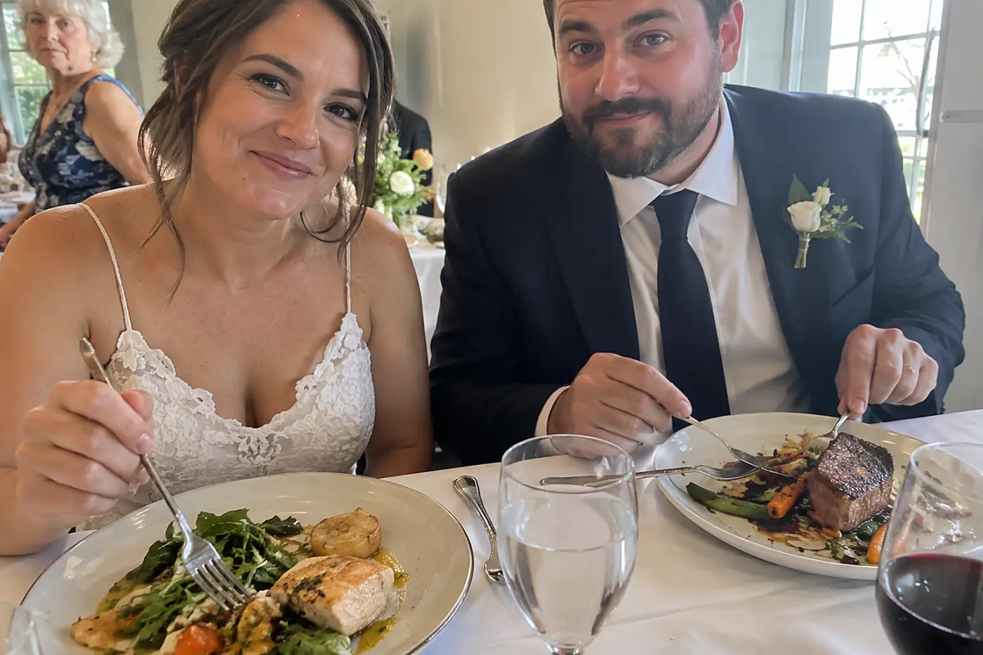 The couple sit with dinner plates at their wedding table while a woman in her sixties passes by without stopping.