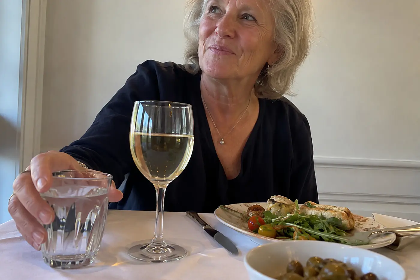 A glass of wine and a glass of water sit before a woman in her sixties at a reception table.