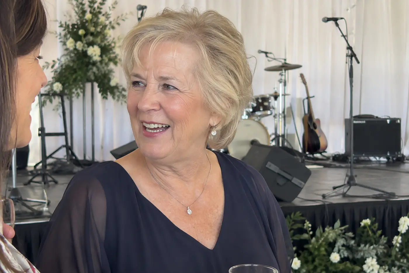 A microphone stands beside a wedding band as a woman in her sixties speaks with family away from the stage.
