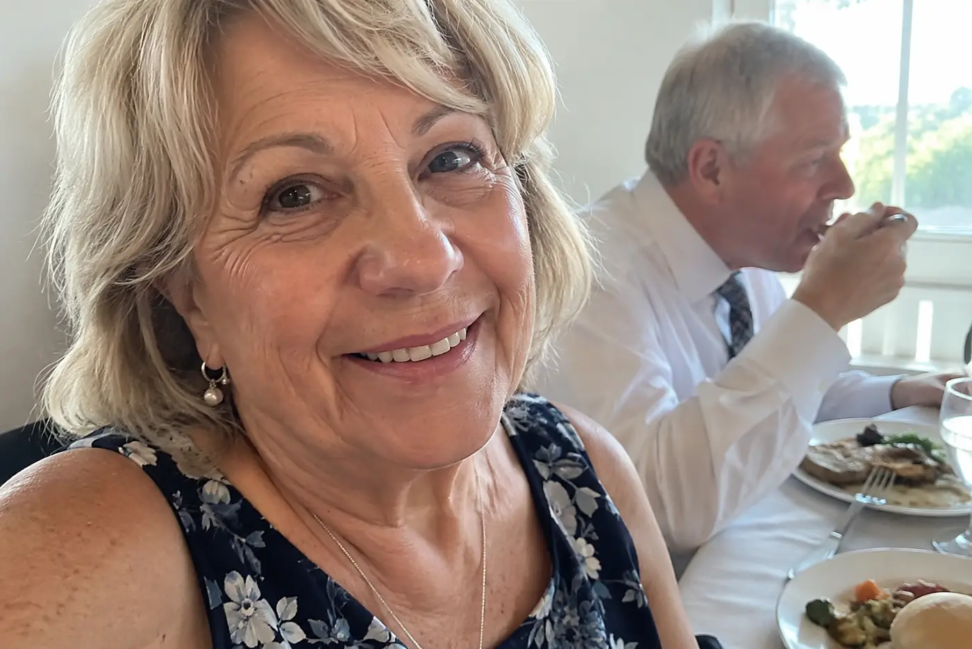 A woman in her sixties lowers her camera at a reception table while another guest eats dinner.