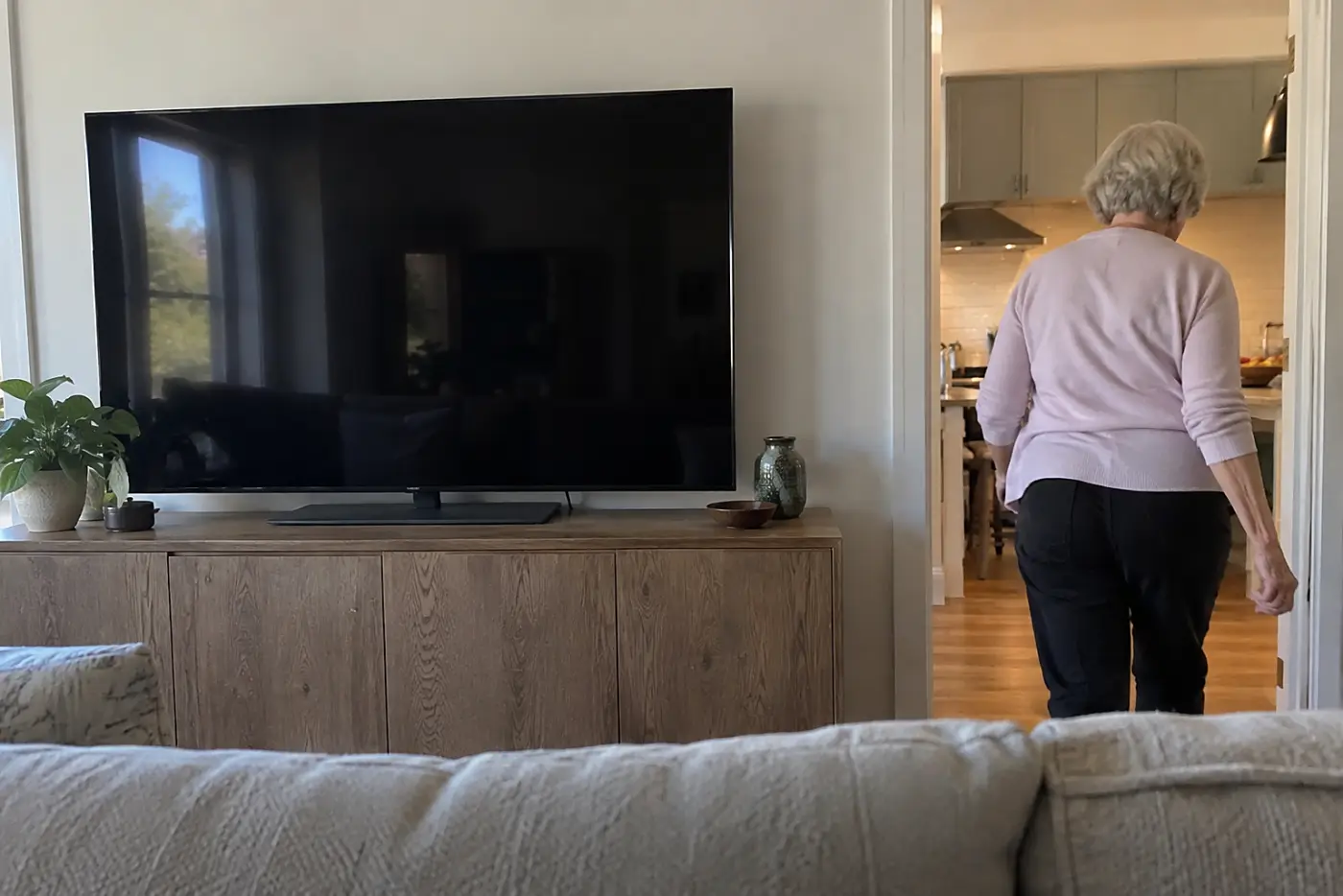 A woman in a pink sweater walks into a kitchen past a large dark television.