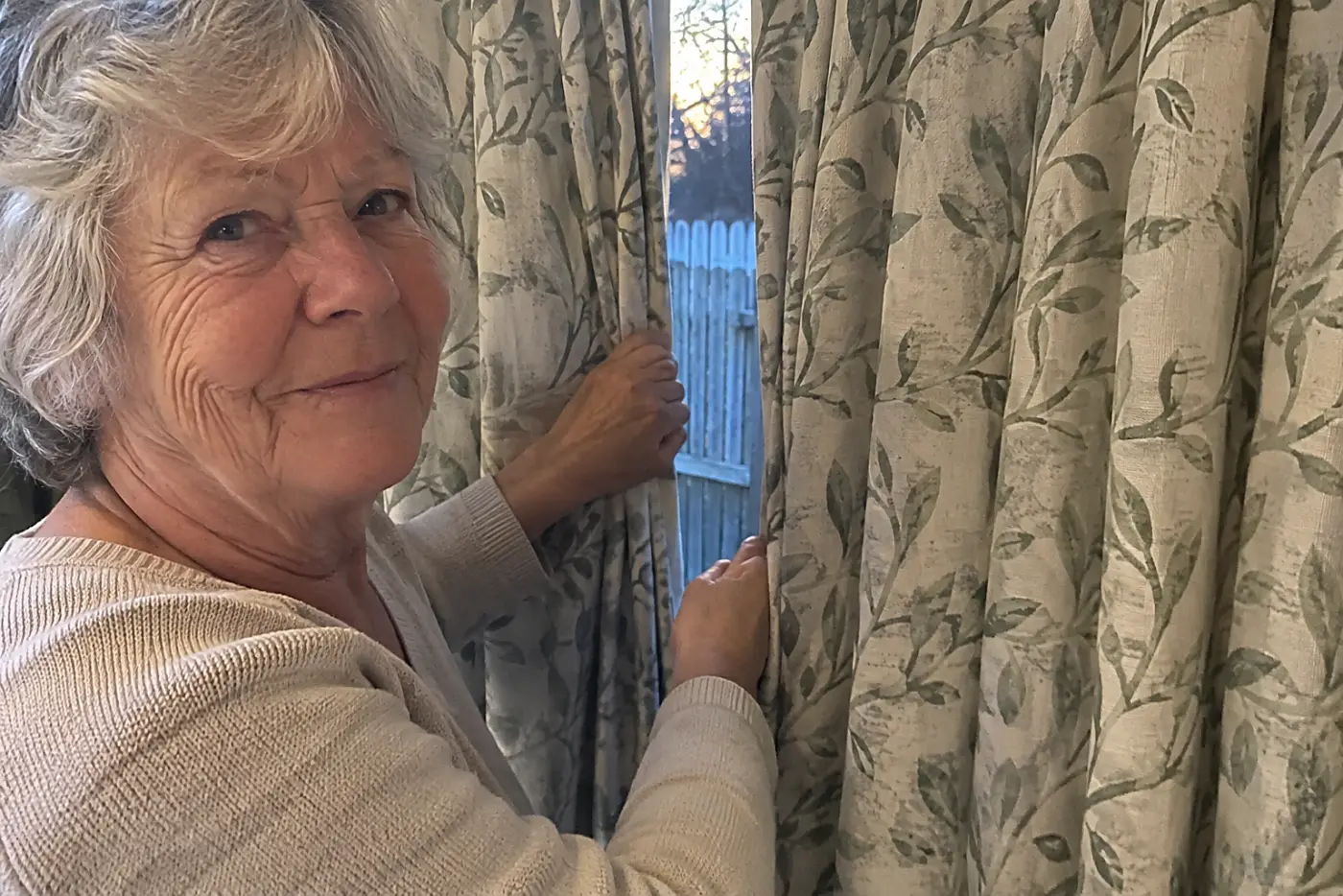 A woman in her seventies pulls patterned curtains closed across a living room window.