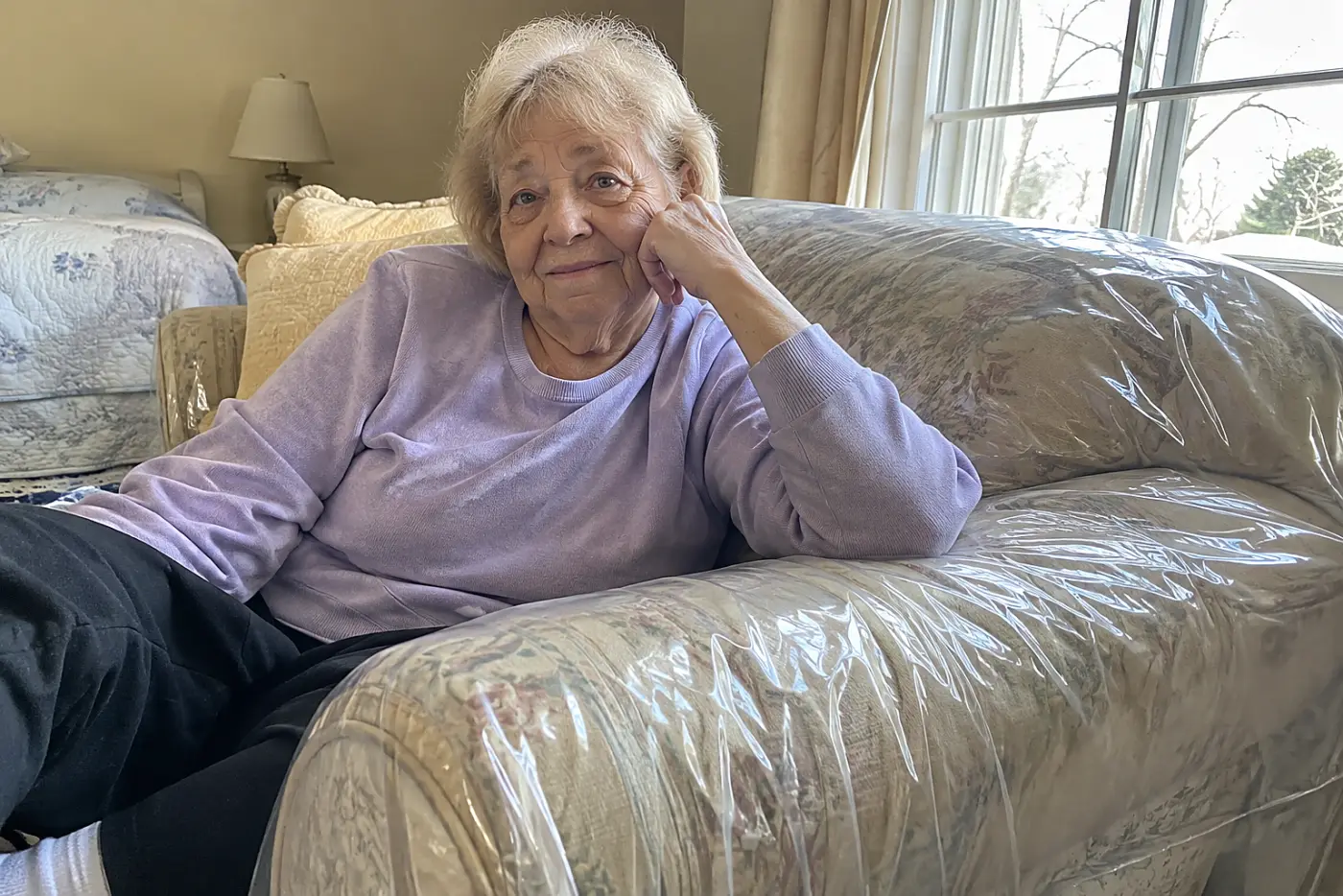 A woman in a purple sweater sits on a sofa wrapped in clear plastic.