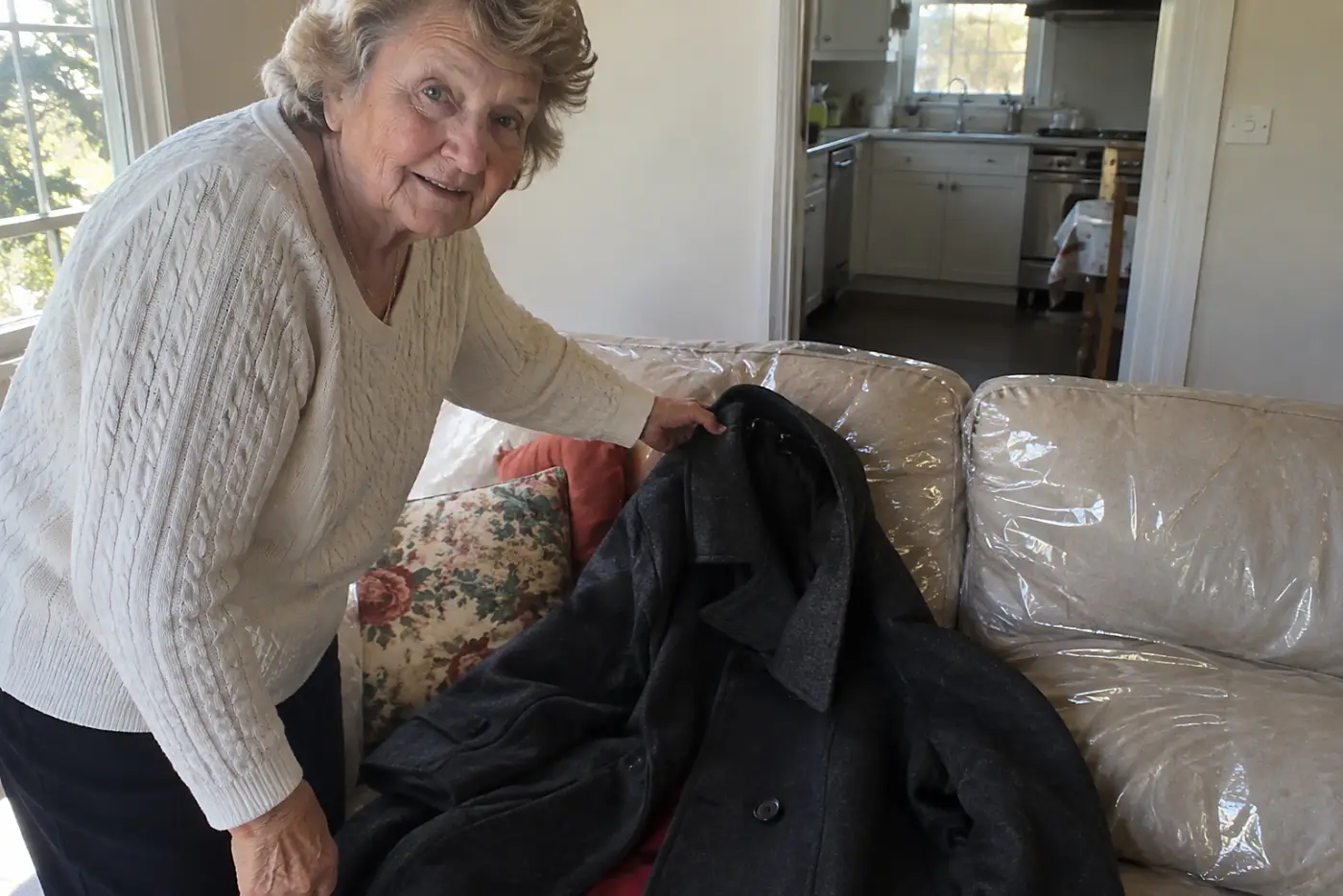 A woman in her seventies lifts a dark coat from a plastic-covered sofa.