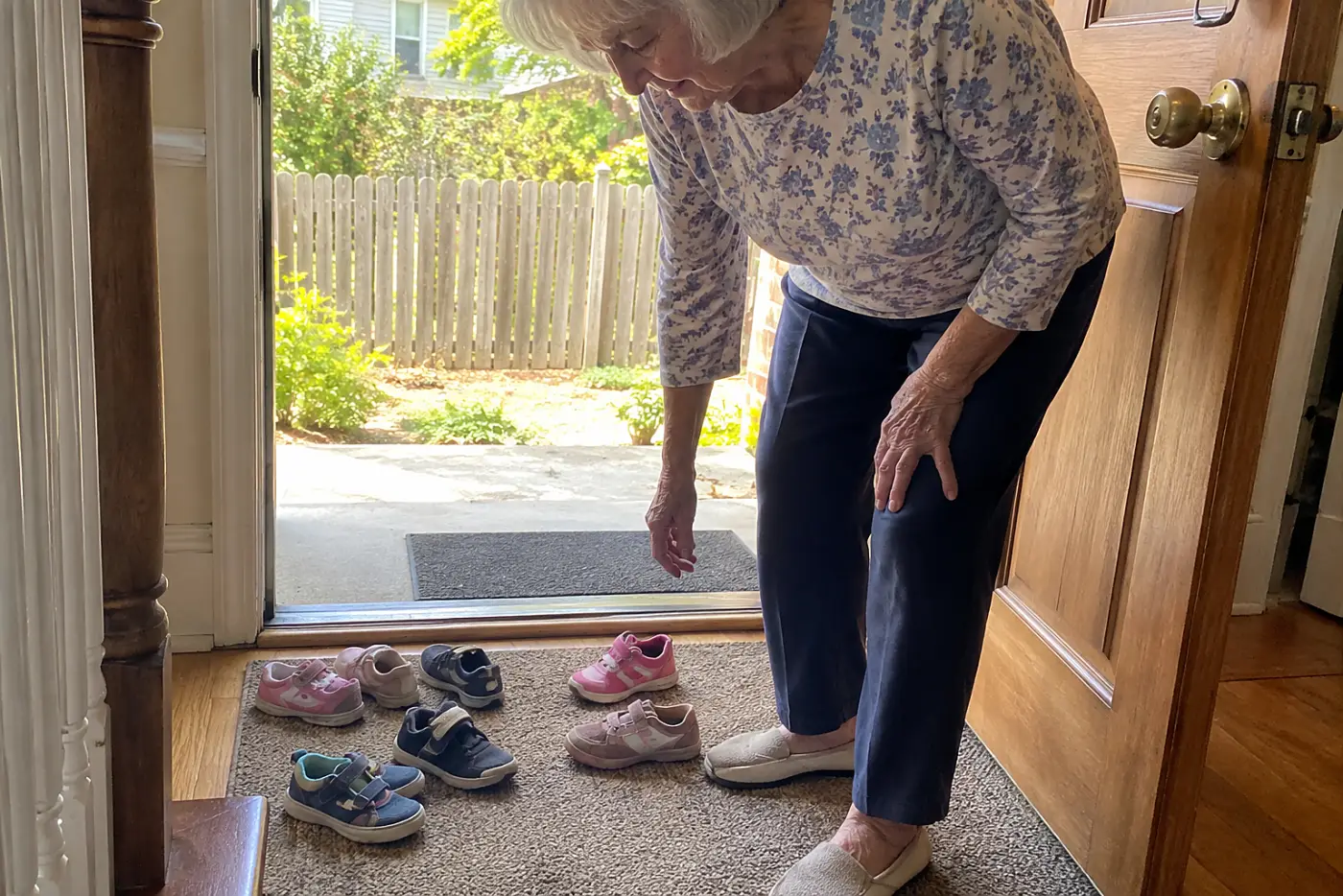 A woman in her seventies reaches down toward small children's shoes on a front mat.