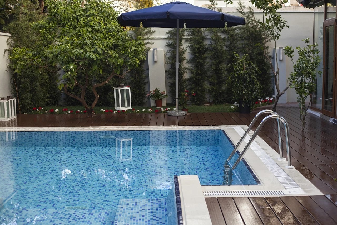 Backyard tiled swimming pool with metal ladder, wooden deck, navy umbrella, lanterns, citrus trees and red flowers