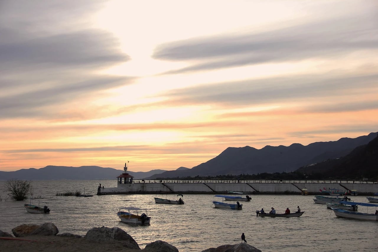 Sunset over Lake Chapala with small fishing boats near a pier and lighthouse, mountains beyond
