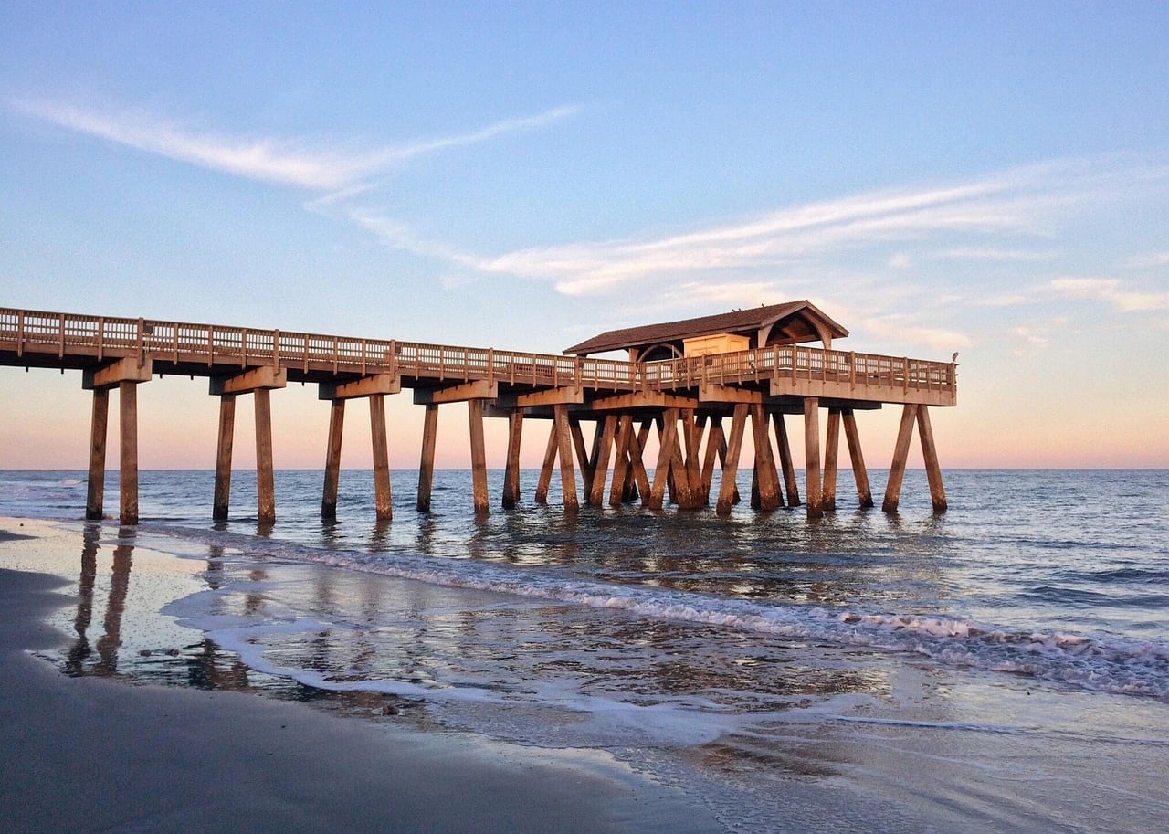 Wooden fishing pier with covered pavilion extending into the ocean at sunset, with gentle waves on the sandy beach