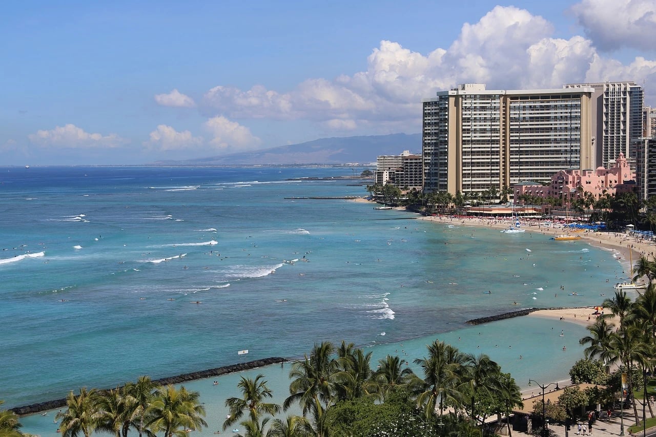 Aerial view of Waikiki Beach with turquoise water, palm trees, high-rise hotels and the pink Royal Hawaiian hotel