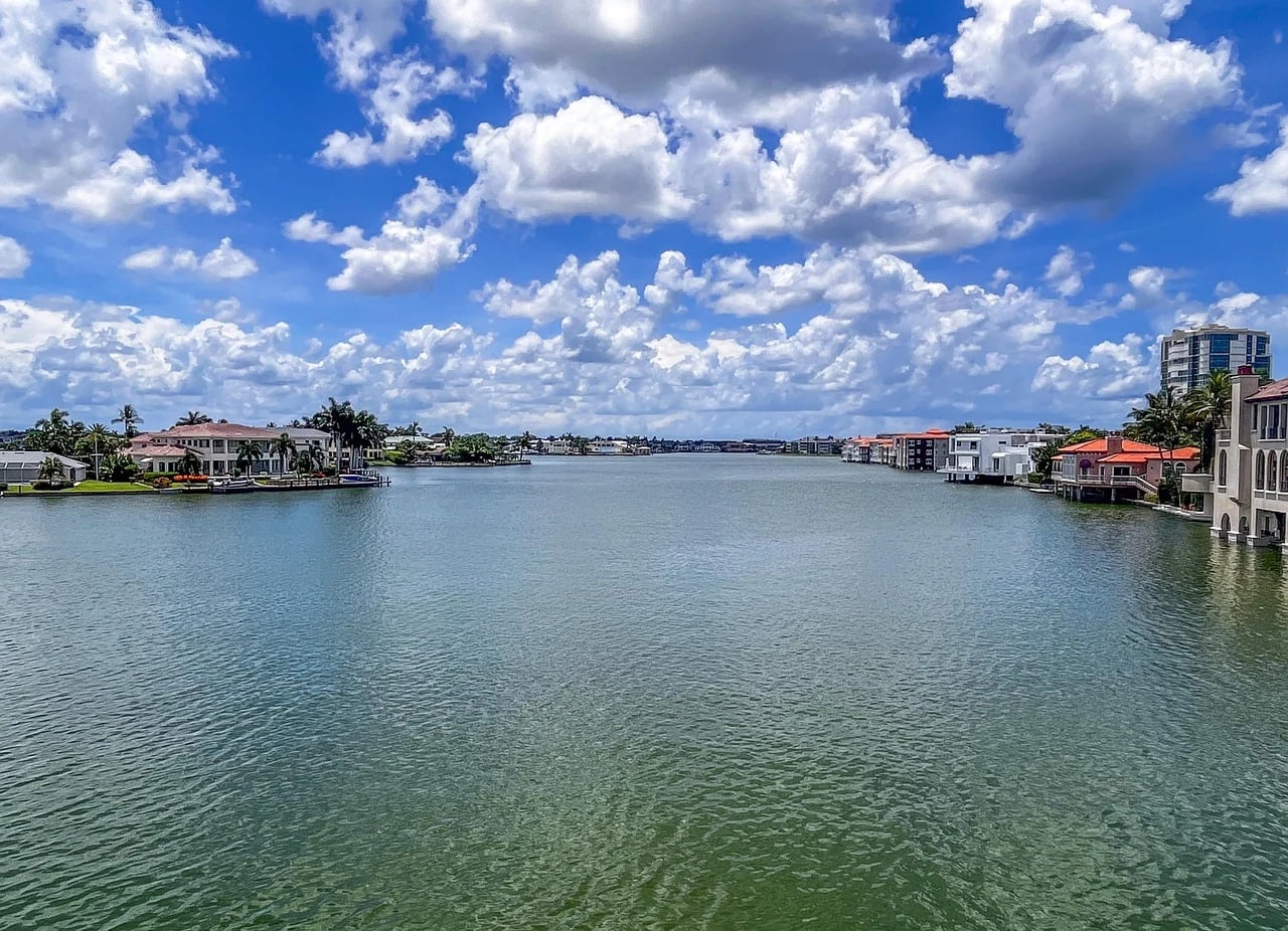 Wide sunny canal lined with palm trees and red-roofed waterfront homes under a blue sky with clouds