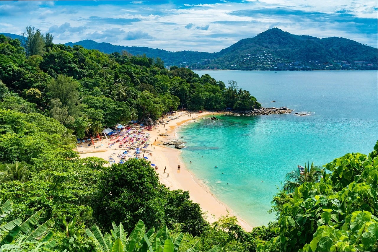 Aerial view of a sandy tropical beach cove with turquoise water, umbrellas and green forested hills in Phuket
