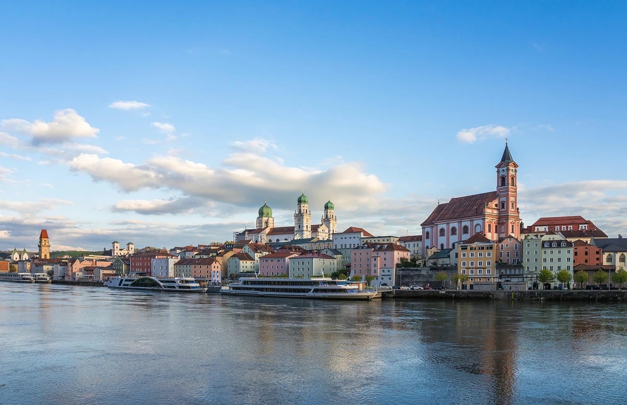 River cruise ships docked along a European riverfront with colorful historic buildings and church towers under a blue sky