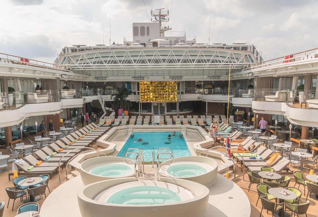 Open pool deck of a mid-sized cruise ship with hot tubs, loungers, tables and a few swimmers
