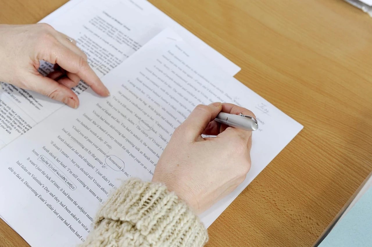 Woman's hands marking edits with a pen on printed manuscript pages lying on a wooden desk