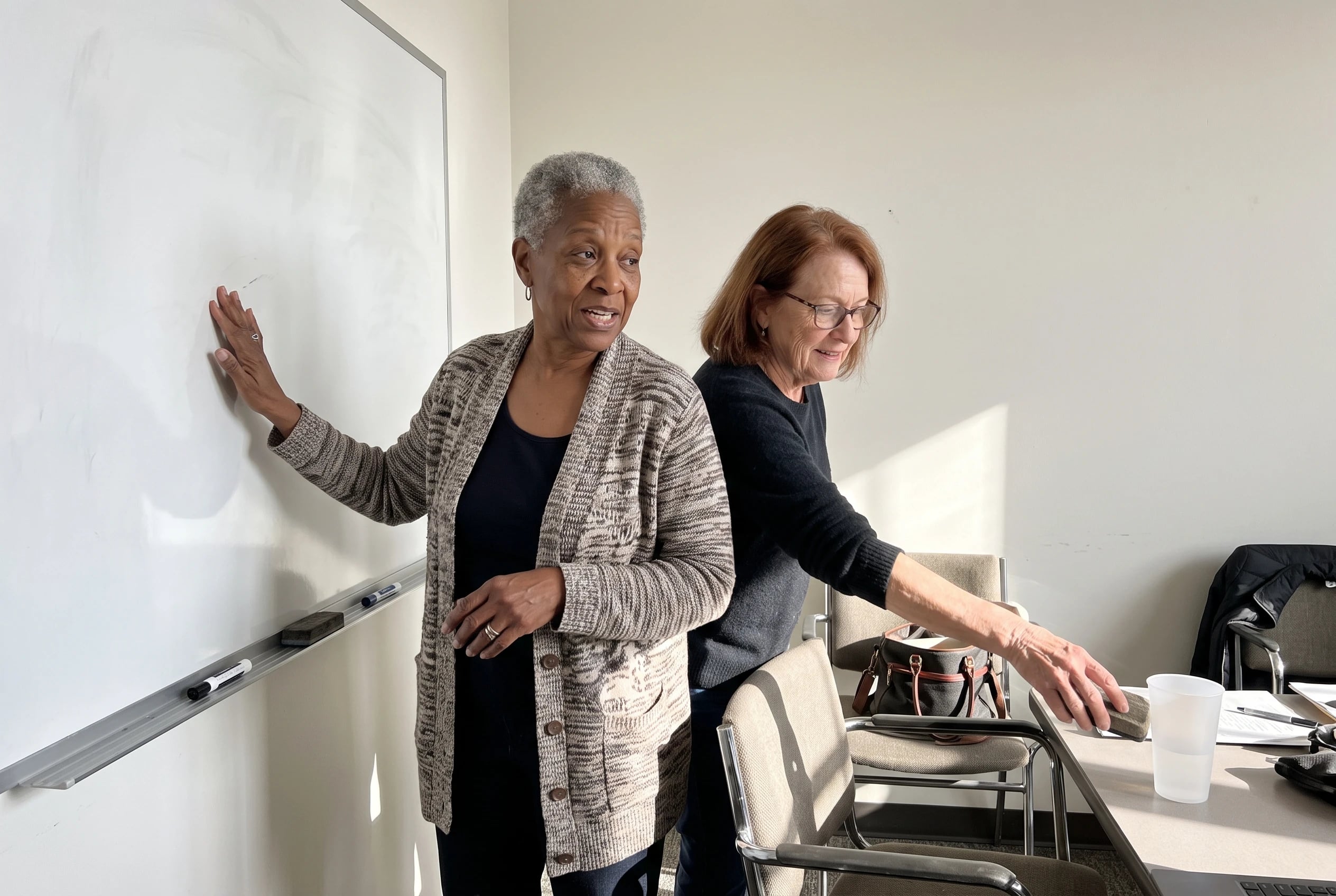 Older woman talking with a former colleague beside a whiteboard in an office meeting room.